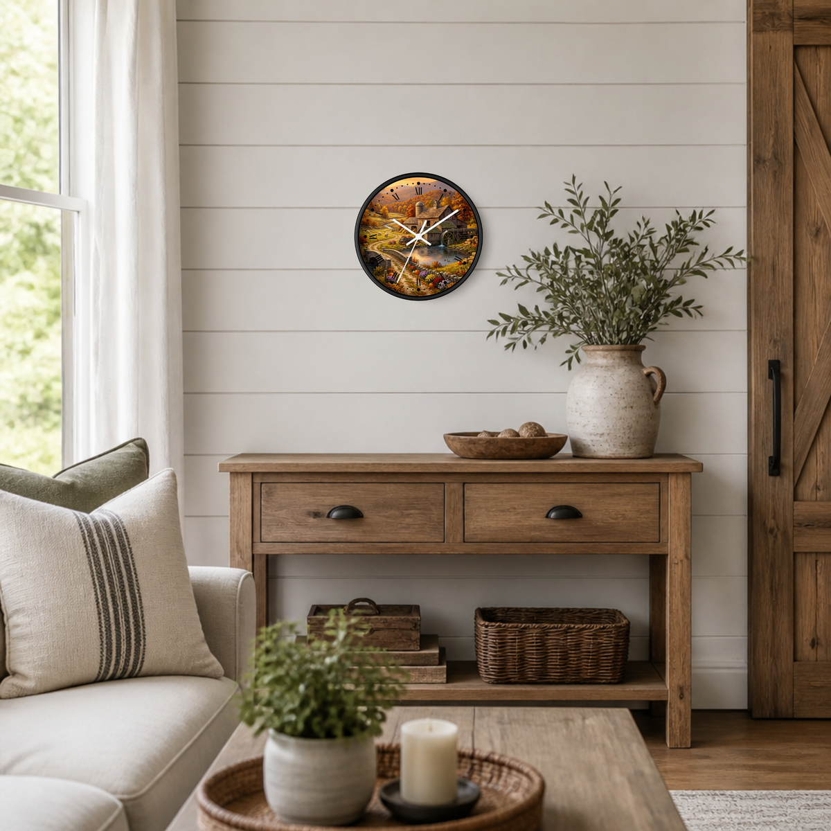 A round wall clock with a rustic autumn farm scene print hangs on a white shiplap wall above a wooden console table in a styled living room.