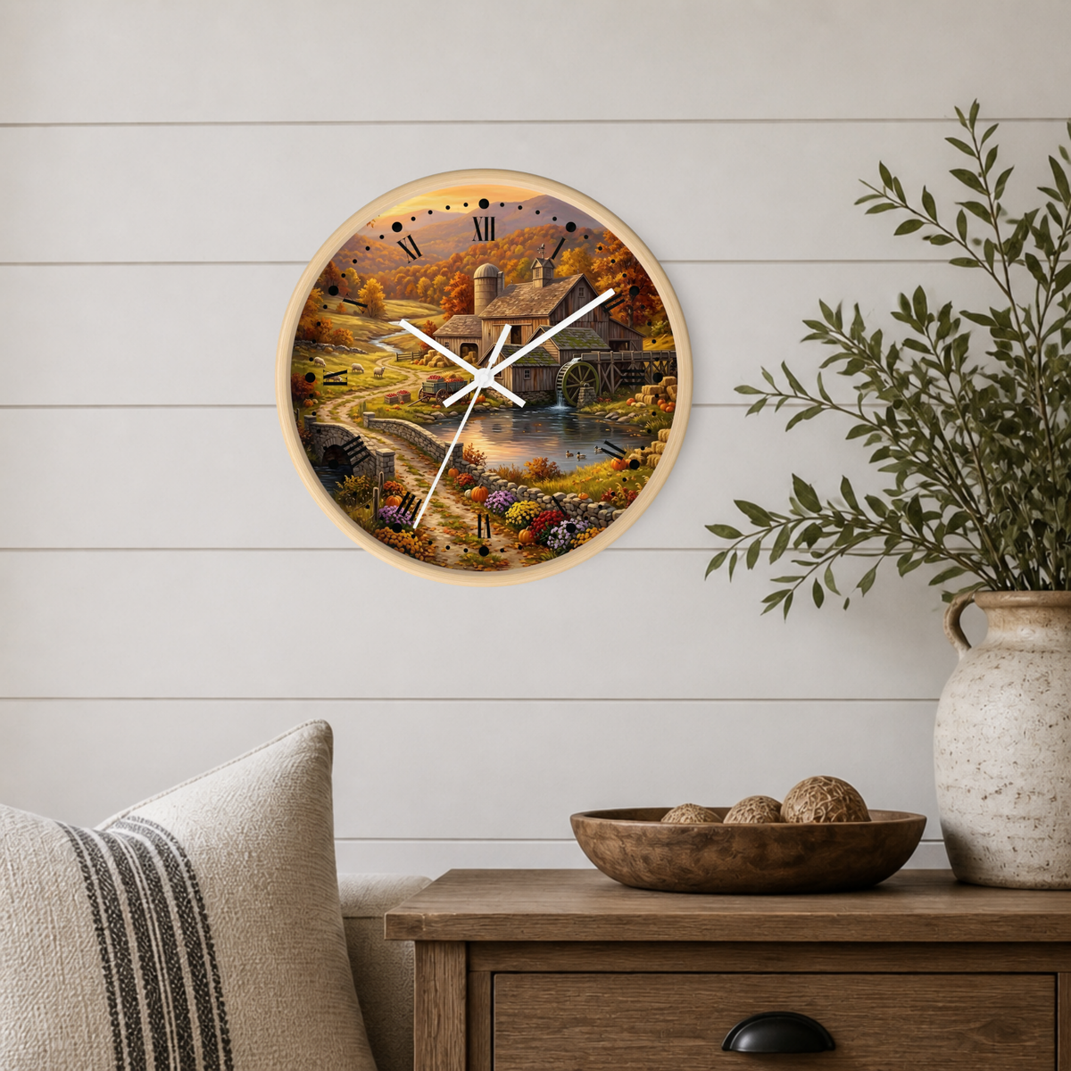 A round wall clock with a light wood frame and a detailed autumn landscape print, mounted on a white shiplap wall above a wooden side table with a vase and bowl.
