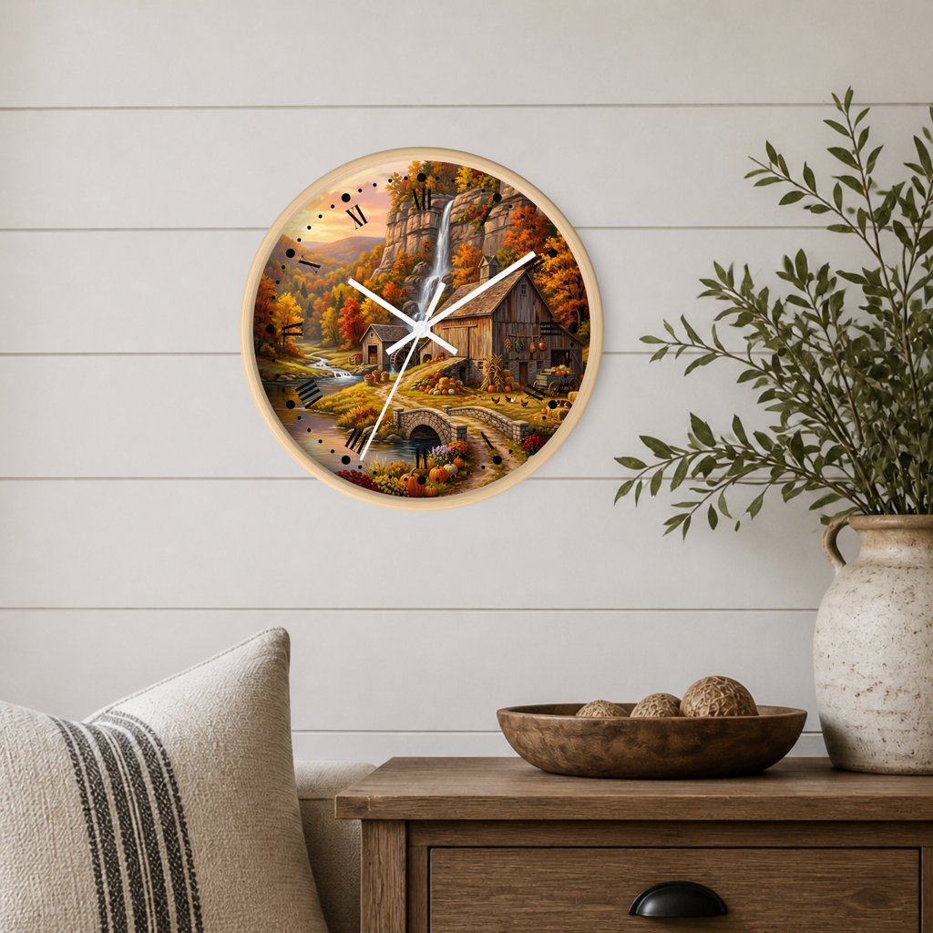 A round wall clock with a light wood frame hangs on a white shiplap wall above a wooden side table. The clock face features a colorful autumn landscape painting with a waterfall, cabin, and fall foliage.