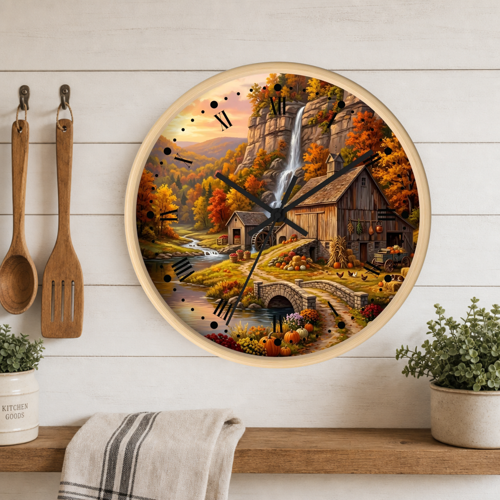 A round wall clock with a light wood frame hangs on a white shiplap wall above a wooden shelf. The clock face features a colorful autumn landscape painting with a waterfall, rustic cabin, bridge, and fall foliage.
