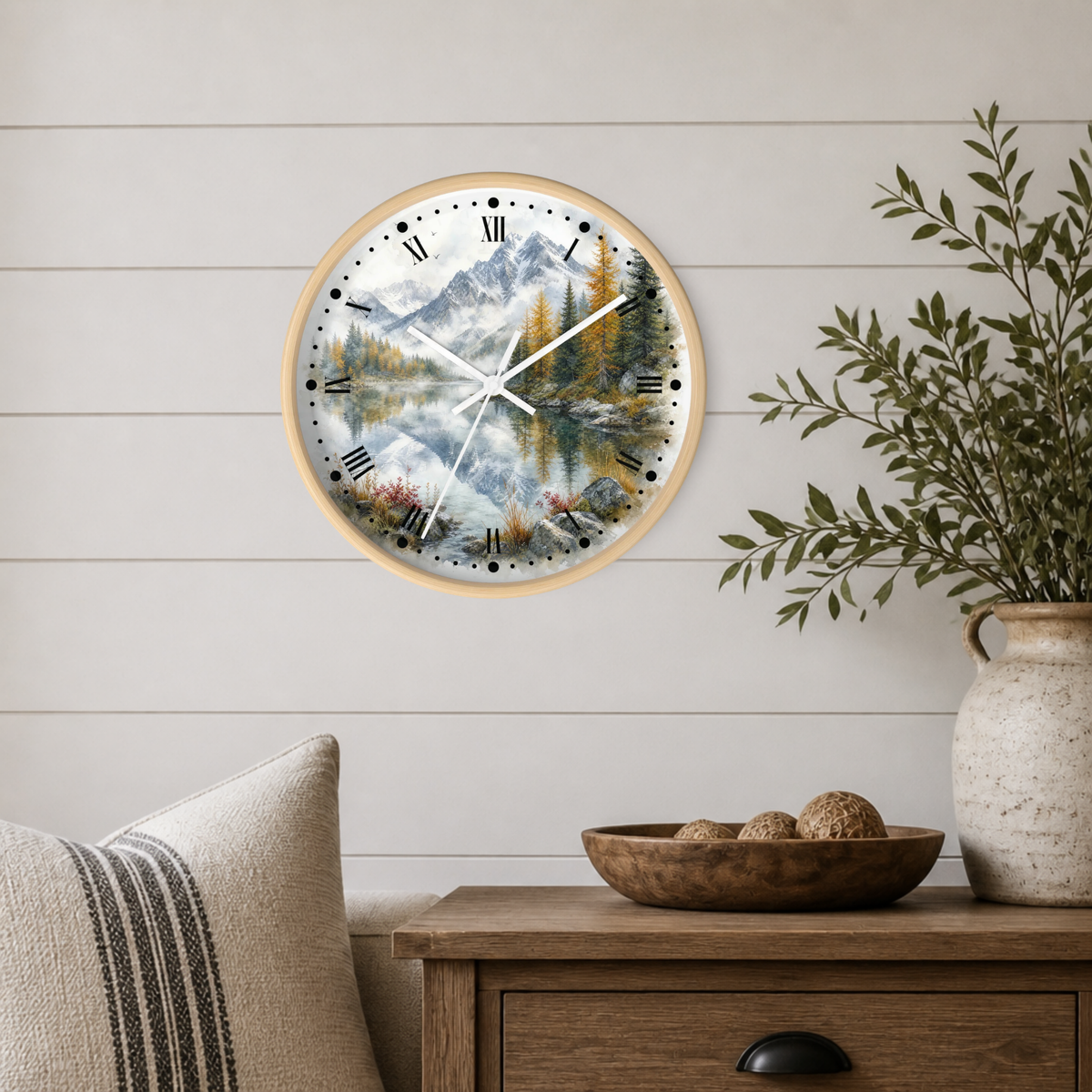 A round wall clock with a light wood frame and a scenic mountain lake print, mounted on a white shiplap wall above a wooden side table with a vase and bowl.