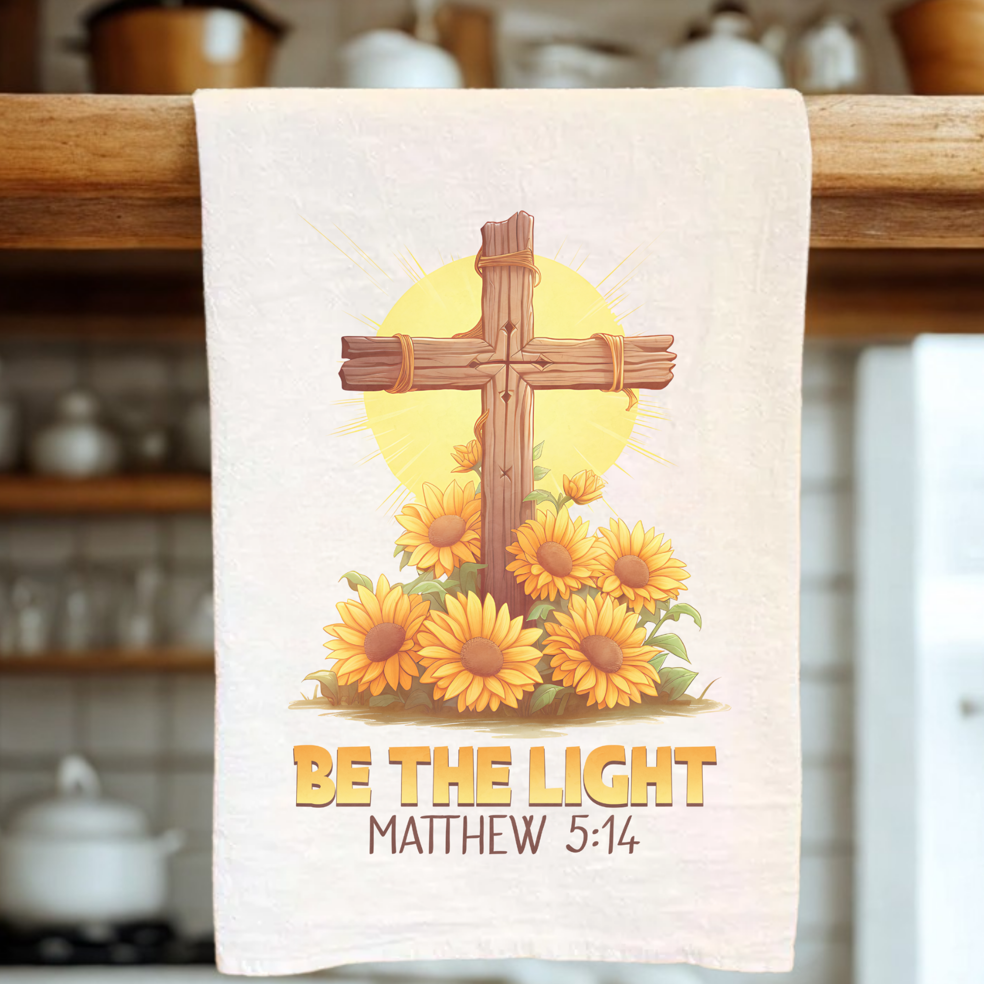 Tea towel with cross and sunflowers design, featuring the text 'Be the Light Matthew 5:14', hanging on a wooden rack.