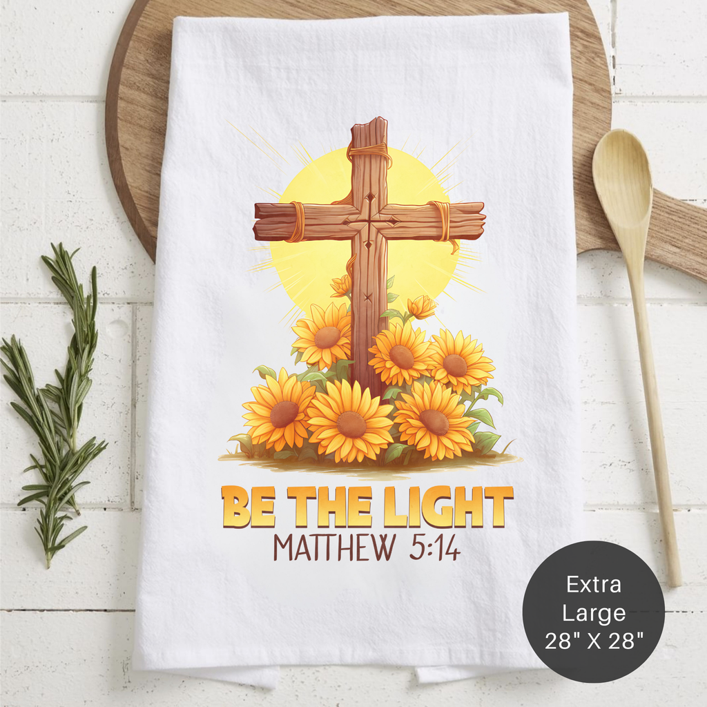 White tea towel with a cross and sunflower design, featuring the text 'Be the Light Matthew 5:14' on a wooden surface.