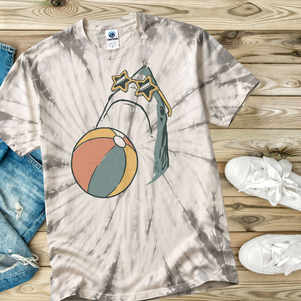 Tie-dye t-shirt in the color Spider Silver with a hand drawn shark with a beach ball and wearing star shaped sunglasses design on a wooden surface with jeans and shoes.