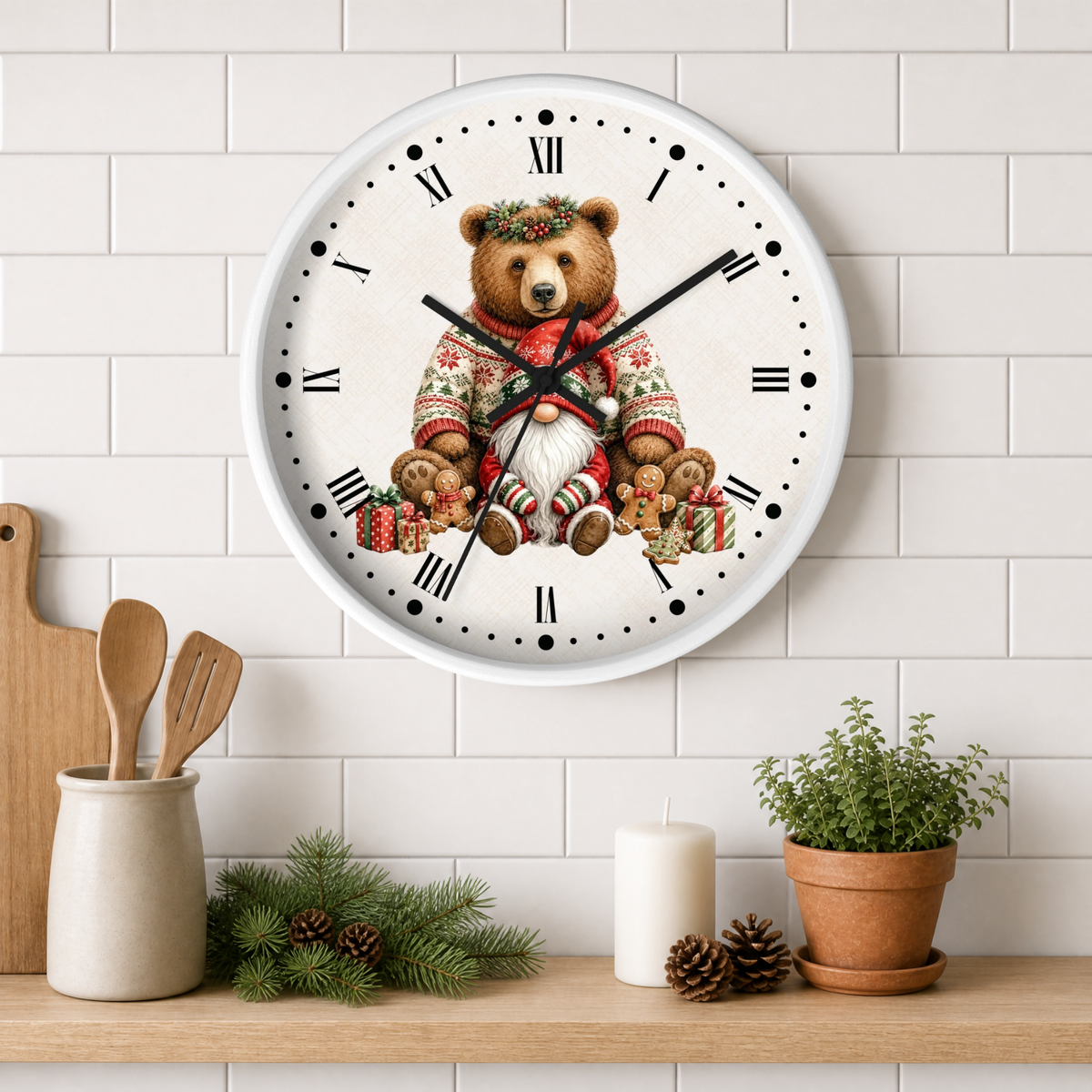 A round wall clock with a white face and Roman numerals, featuring a teddy bear in a red Christmas sweater and green wreath. The clock is mounted on a white subway tile wall above a wooden shelf styled with kitchen utensils, pine branches, a candle, and a potted plant.
