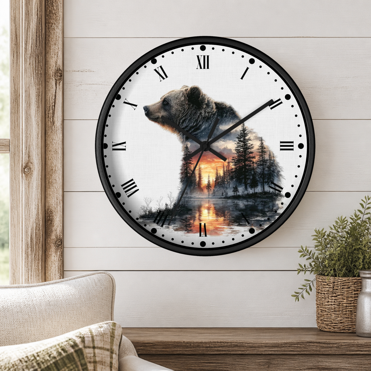 A round wall clock with a black frame and a printed design of a bear over a forest and lake at sunset, mounted on a white wood-paneled wall above a wooden shelf with a plant and part of a chair visible.