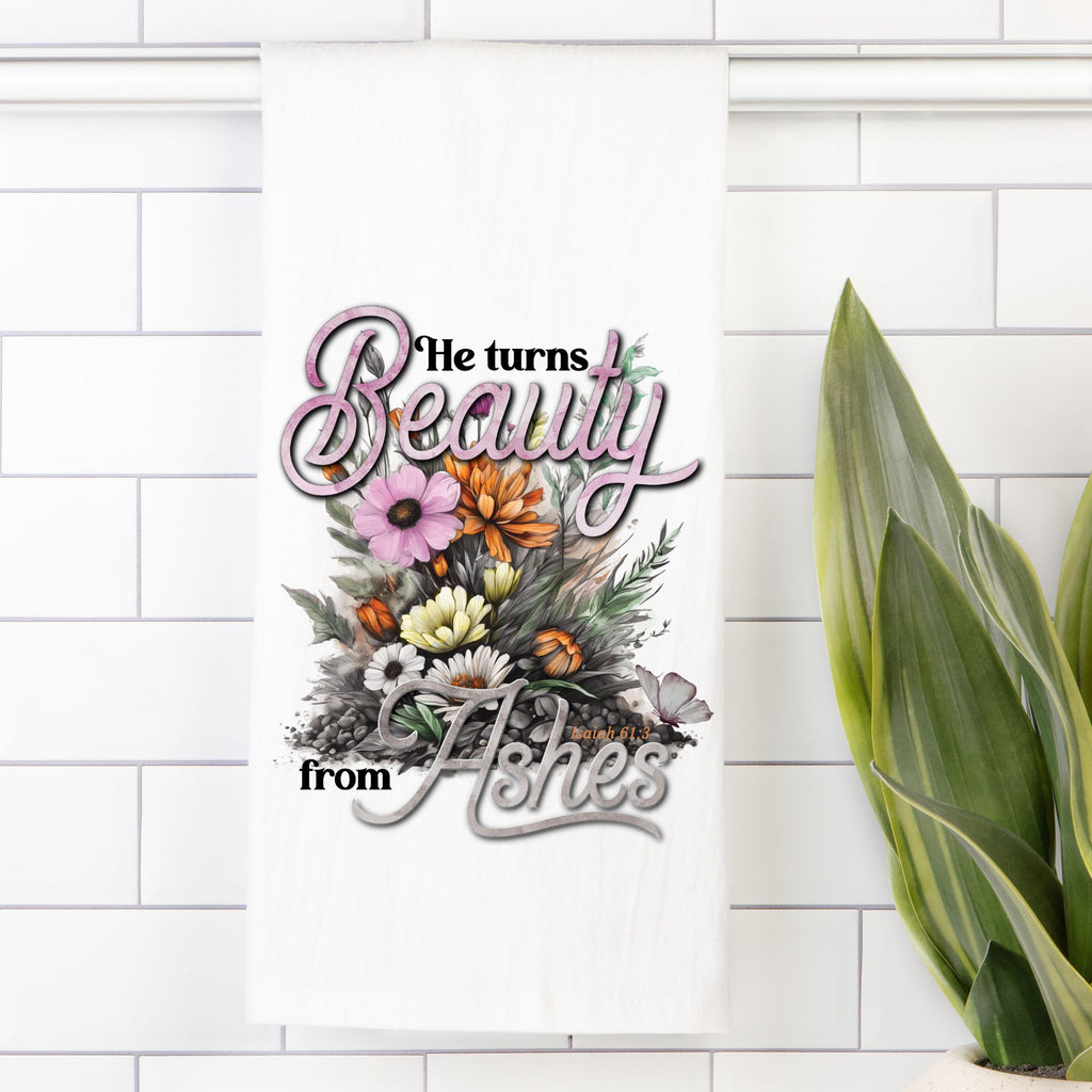 Decorative tea towel with floral design and text 'He turns Beauty from Ashes' on a white tiled wall.