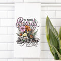 Decorative tea towel with floral design and text 'He turns Beauty from Ashes' on a white tiled wall.