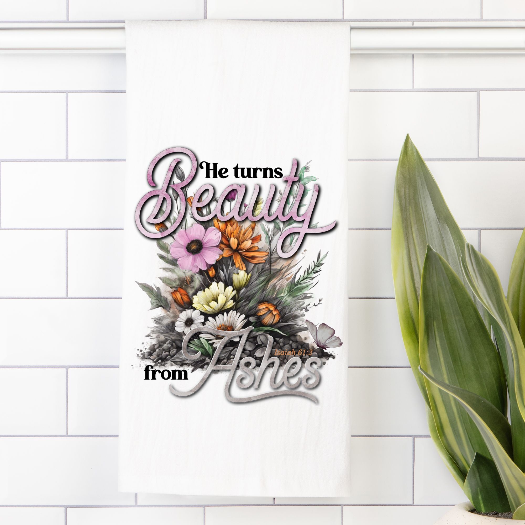 Decorative tea towel with floral design and text 'He turns Beauty from Ashes' on a white tiled wall.