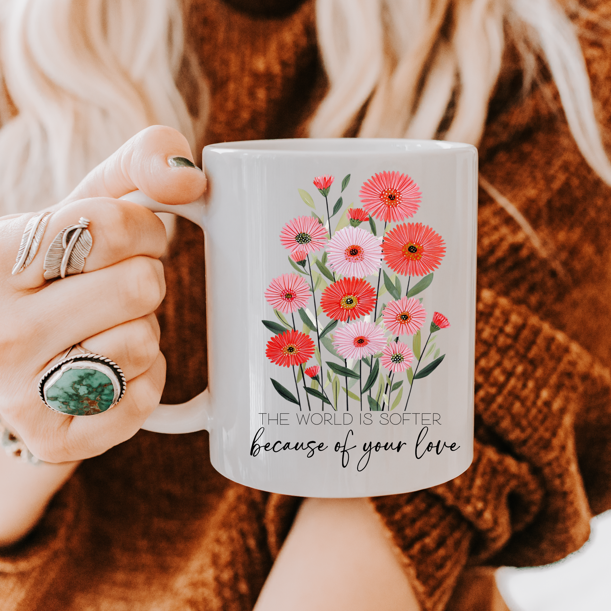 Person holding an 11oz mug with floral design and text 'The world is softer because of your love', wearing a brown sweater.