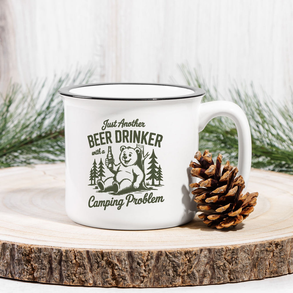 White mug with bear graphic and text on a wooden surface with a pine cone and evergreen branches.