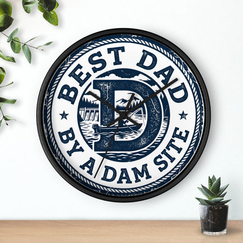 Black wall clock with 'Best Dad by a Dam Site' text on a white wall with a plant and small pot.