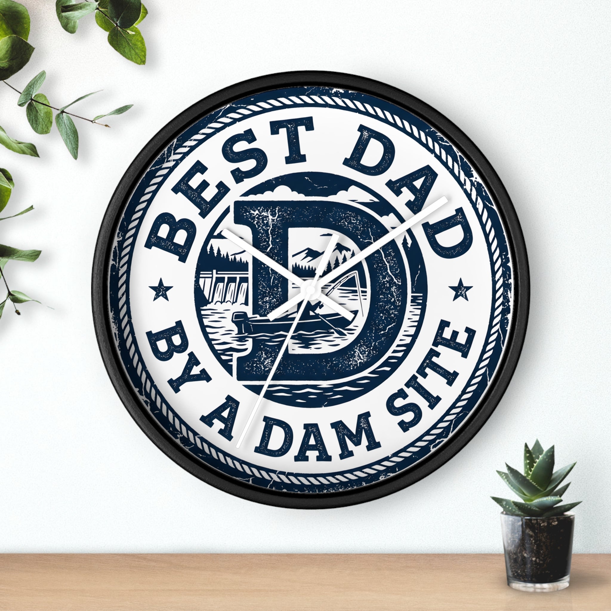 Black Wall clock with 'Best Dad by a Dam Site' text on a white wall with a plant and plant pot.