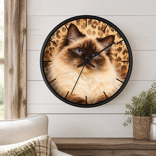 A round wall clock with a black frame and a printed cat face design on the face, mounted on a white shiplap wall above a wooden shelf with a small potted plant.