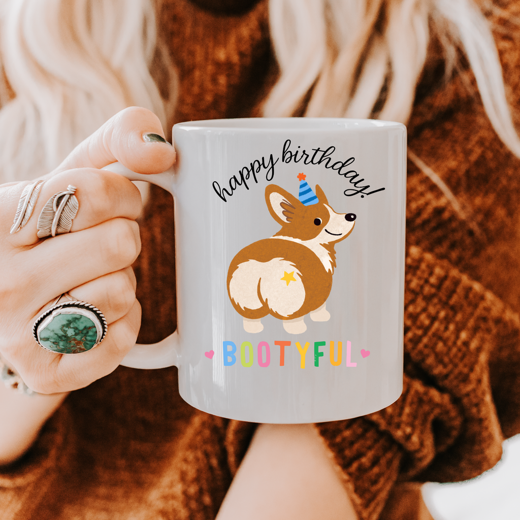 Person holding a mug with a corgi graphic and 'Happy Birthday Bootyful' text.