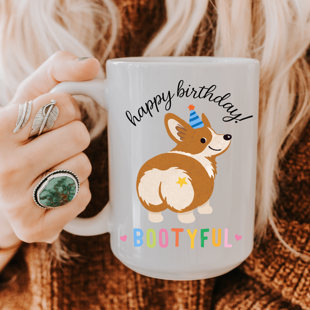 Mug with a corgi design and 'happy birthday! Bootyful' text held by a person.