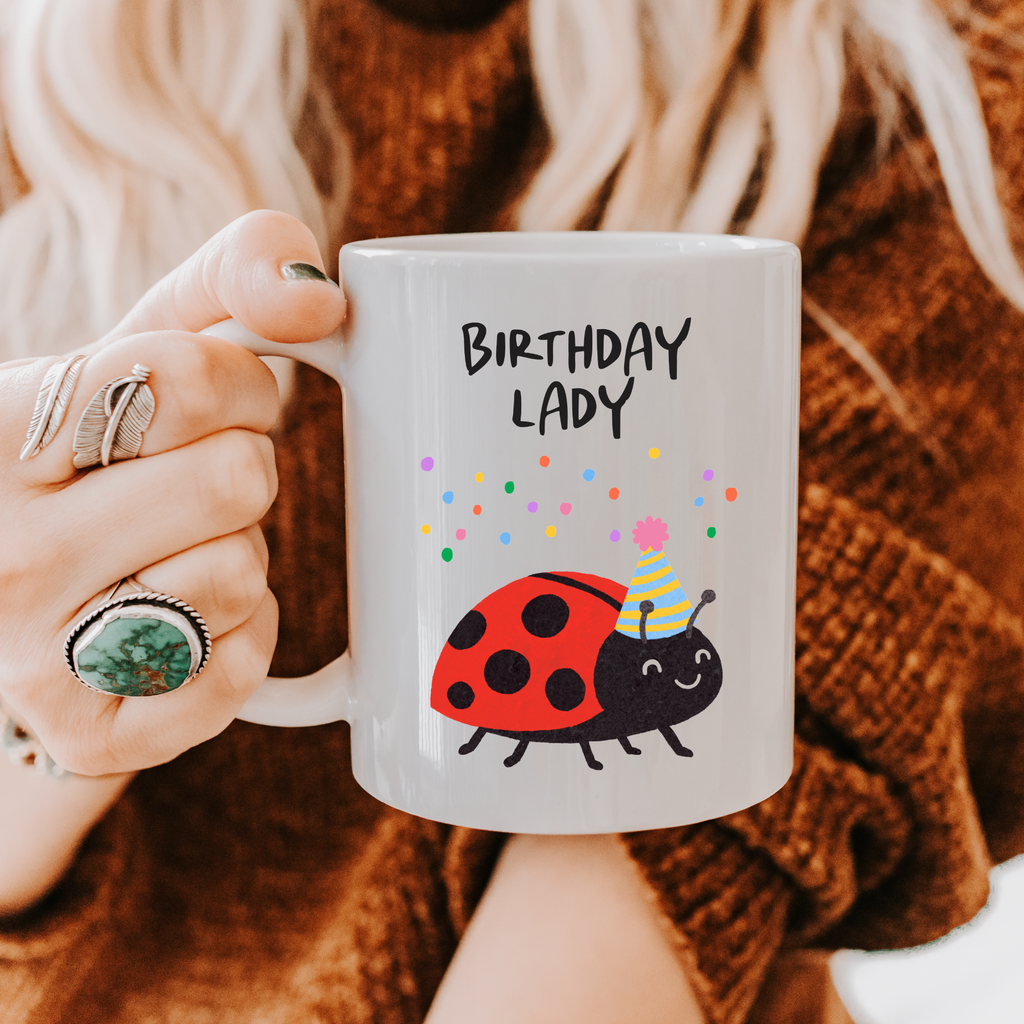 Person holding a mug with 'Birthday Lady' text and ladybug design, wearing a brown sweater.