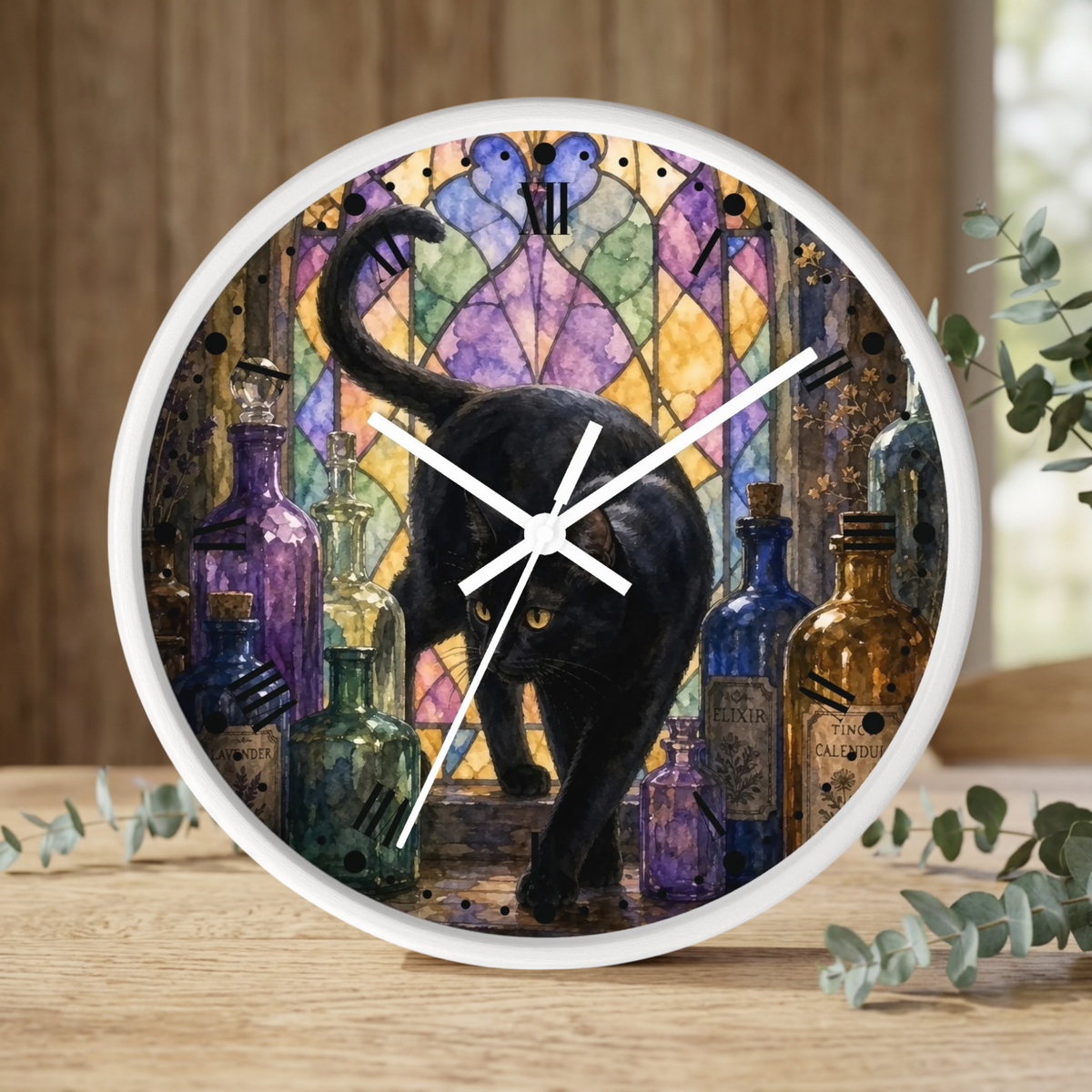 A round wall clock with a white frame and black cat artwork in front of a stained-glass-style background, shown standing on a wooden surface with eucalyptus leaves around it.