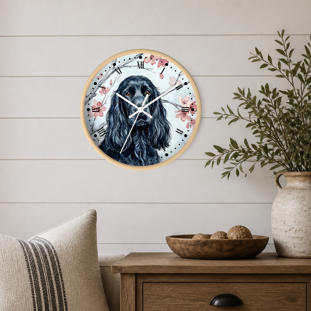 A round wall clock with a black-and-white dog portrait and pink floral details, mounted on a light wood-paneled wall above a wooden side table with a vase and pillows.
