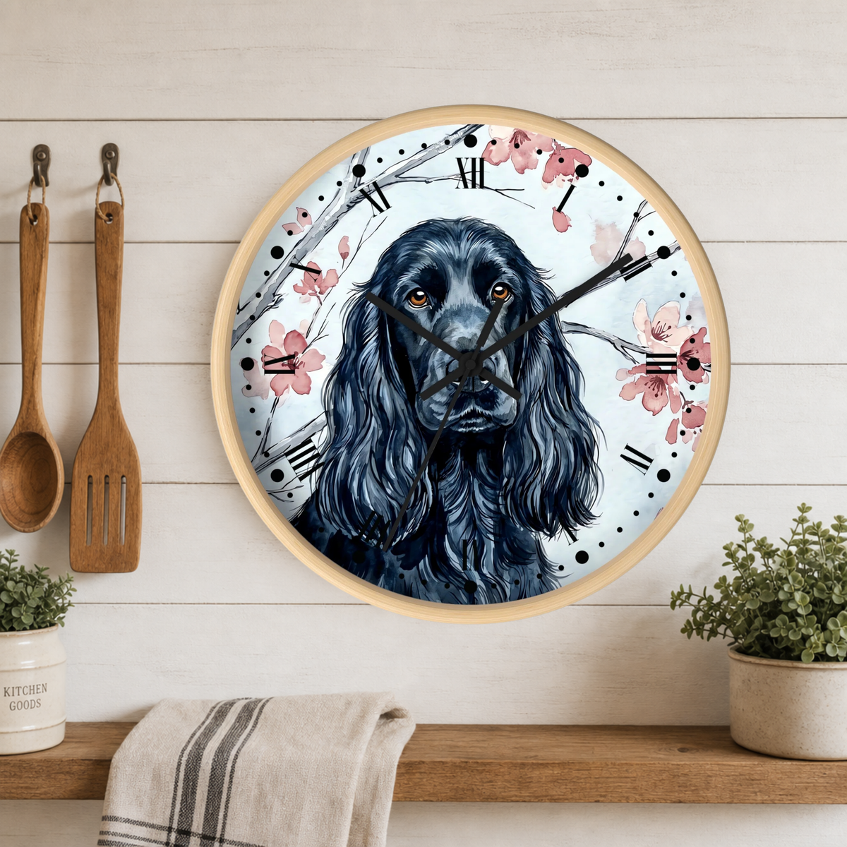 Round wall clock with a light wood frame featuring a black-and-blue dog portrait, pink cherry blossoms, and black Roman numerals, mounted on a white wood plank wall above a shelf with plants and kitchen utensils.