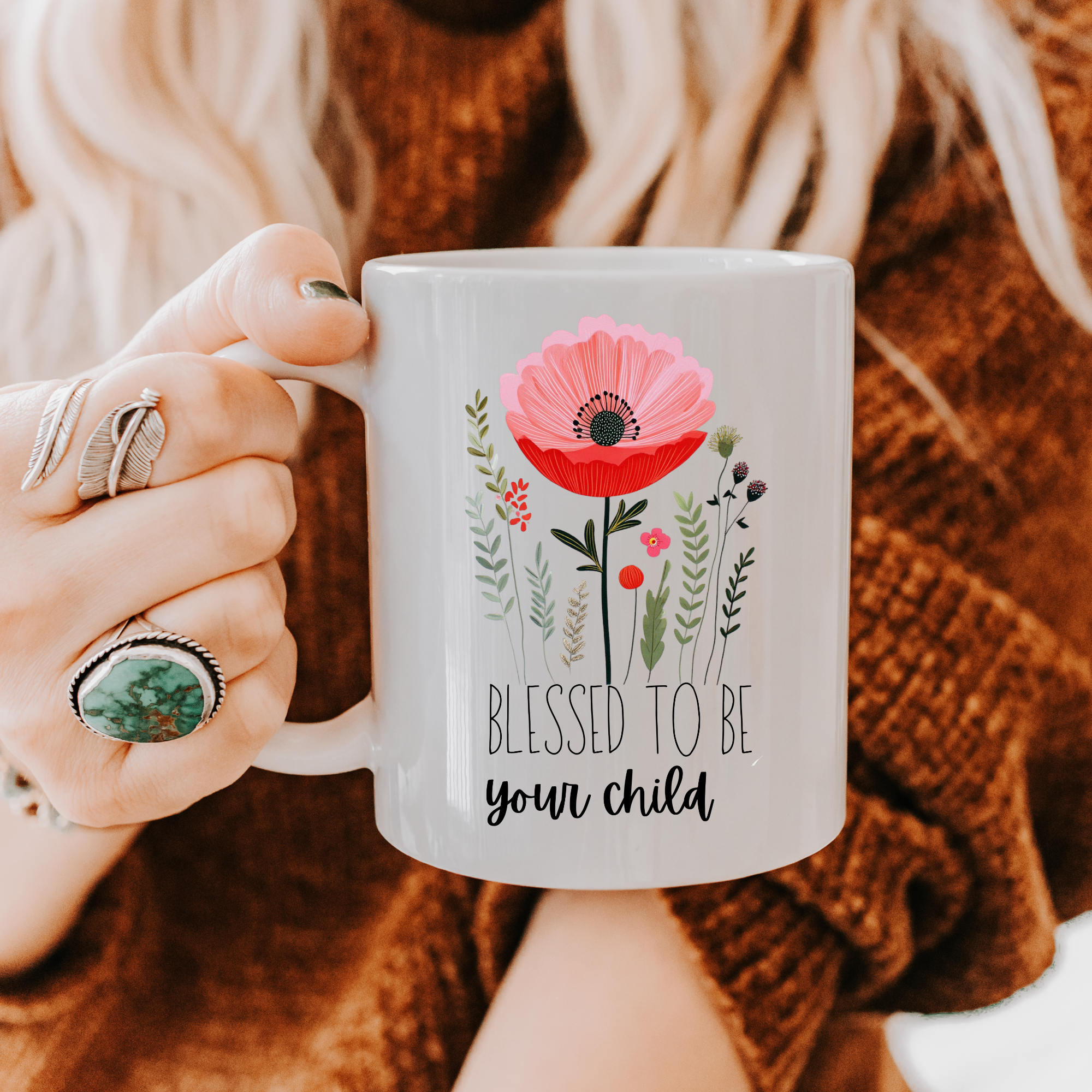 Person holding an 11oz mug with a hand drawn floral design and text 'Blessed to be your child', wearing a brown sweater.