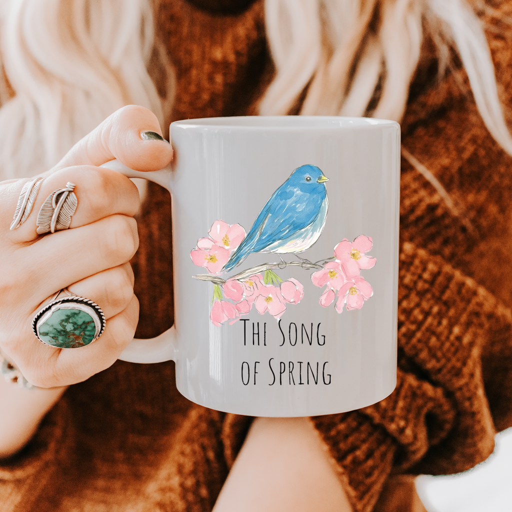 Person holding a mug with a blue bird and pink flowers design, text 'The Song of Spring', wearing a brown sweater.