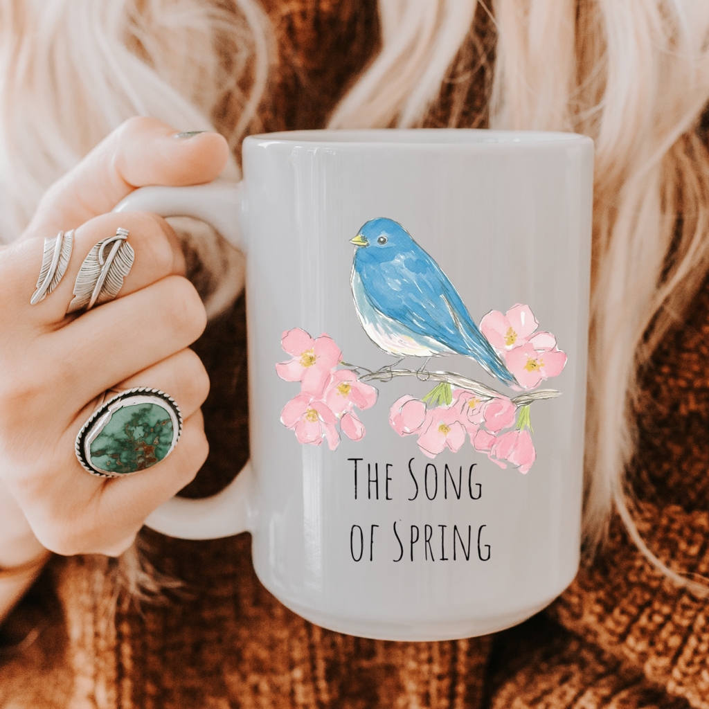 Person holding a mug with a blue bird and cherry blossoms design, text 'The Song of Spring', on a textured fabric background.