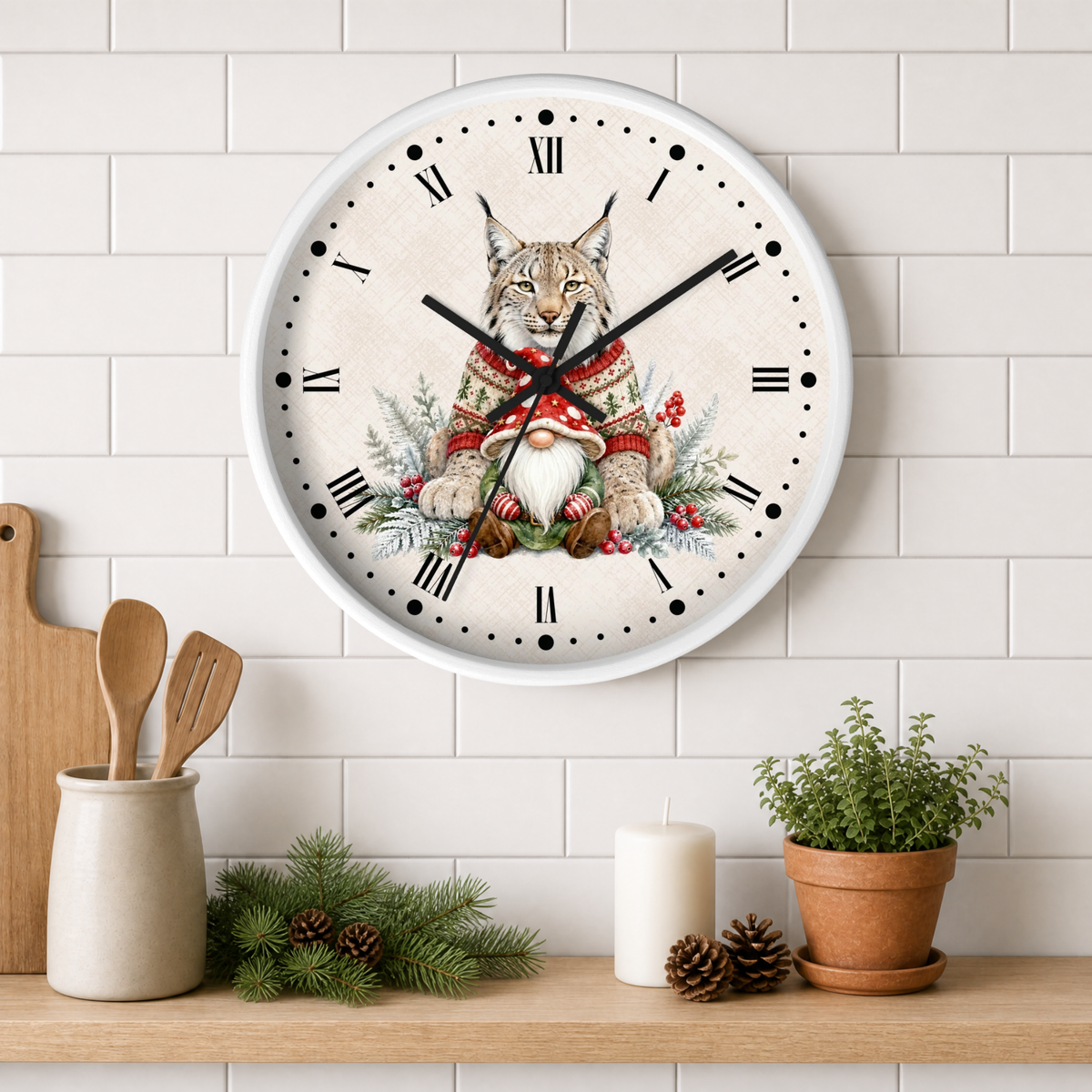 A round wall clock with a white frame and Roman numerals, featuring a festive illustration of a lynx wearing a red knit sweater and a white bearded gnome surrounded by greenery and red berries. The clock is mounted on a white subway tile wall above a wooden shelf styled with kitchen utensils, pine branches, a candle, and a potted plant.