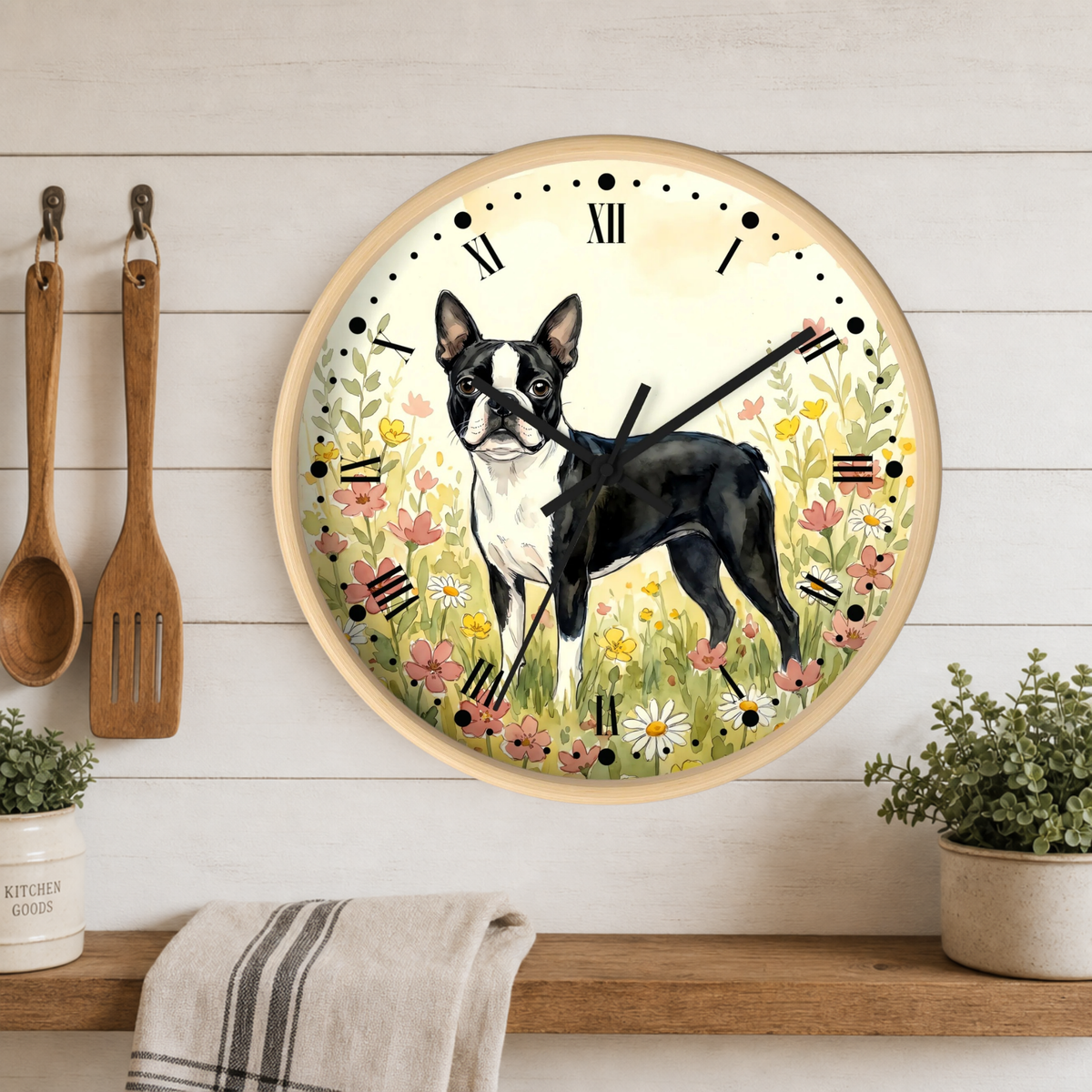 A round wall clock with a light wood frame hangs on a white wood plank wall above a wooden shelf. The clock face features a watercolor-style black and white Boston Terrier standing in a field of pink, yellow, and white flowers.
