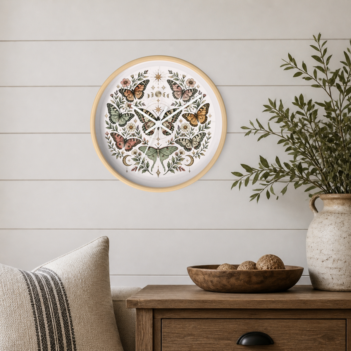Round framed wall art featuring a symmetrical butterfly and floral illustration, displayed on a white shiplap wall above a wooden side table with a vase and bowl.