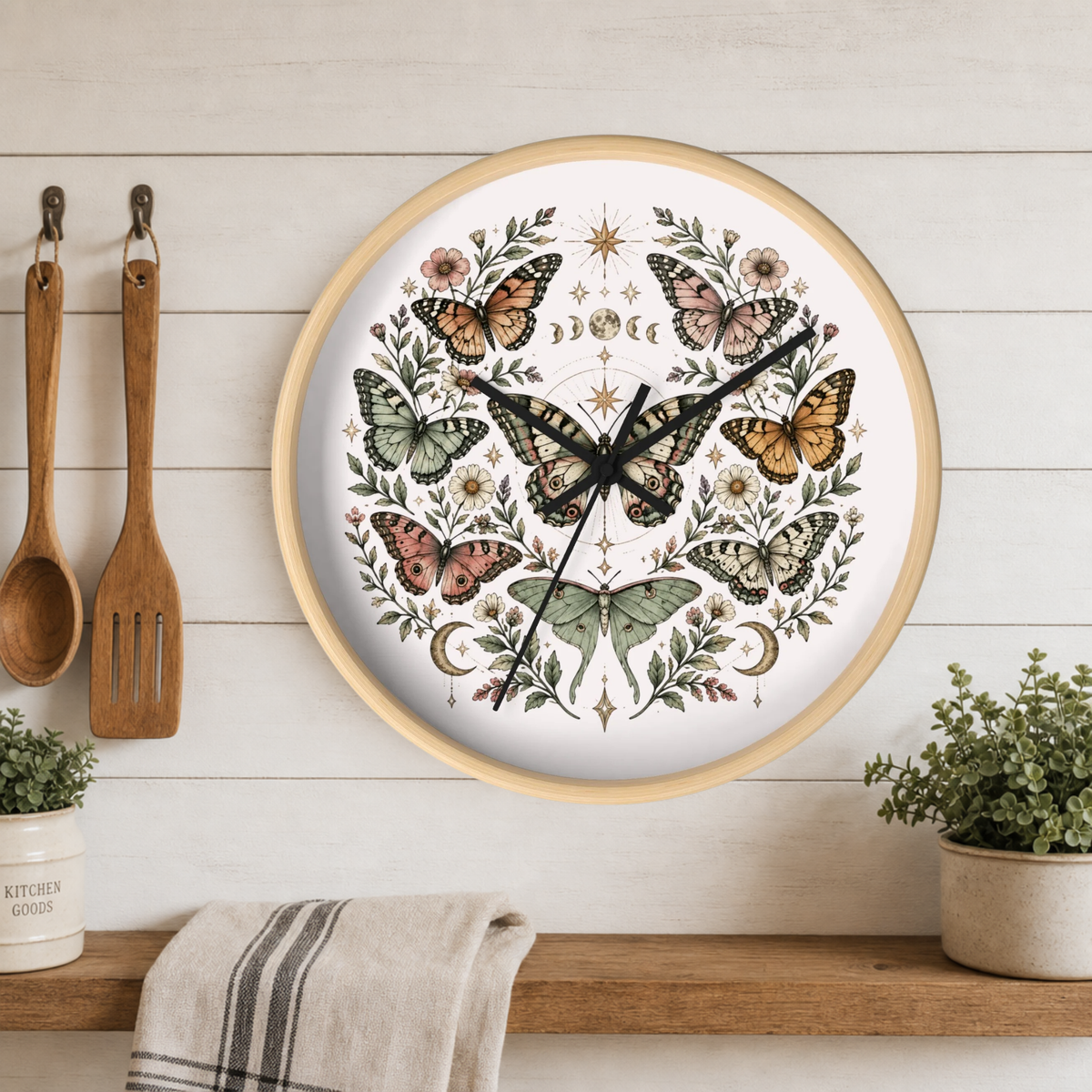 Round wall clock with a light wood frame and a white face decorated with a symmetrical butterfly and floral illustration, hanging on a white wood plank wall above a shelf with plants and kitchen utensils.