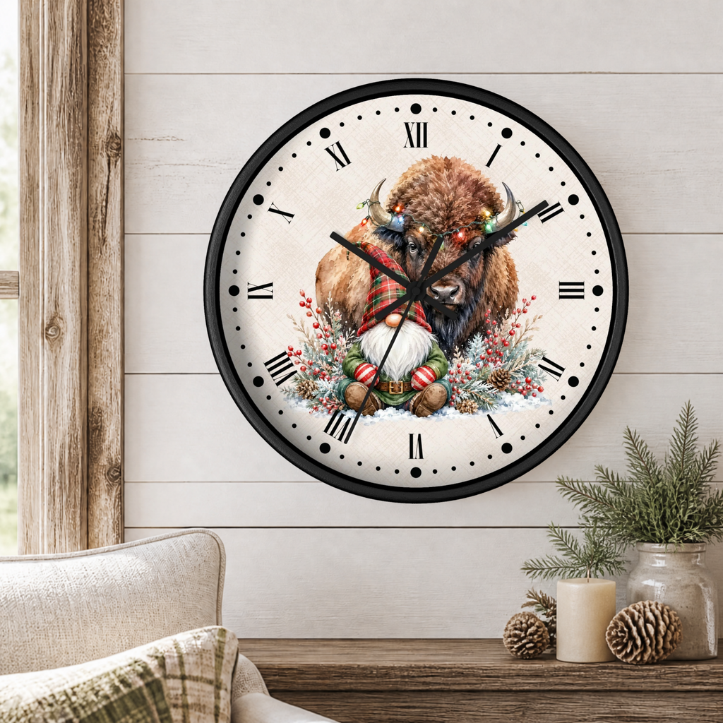 A round wall clock with a black frame and a festive winter illustration of a bison, gnome, wreath, and ornaments on the face, mounted on a white wood paneled wall above a rustic table with candles and pine branches.