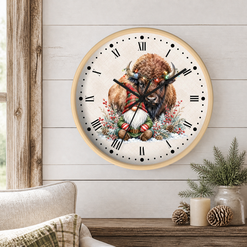 A round wall clock with a light wood frame hangs on a white shiplap wall above a rustic wooden table. The clock face features a detailed illustration of a festive bison wearing a red and green plaid scarf and a red-and-white striped hat, surrounded by a wreath and winter greenery.
