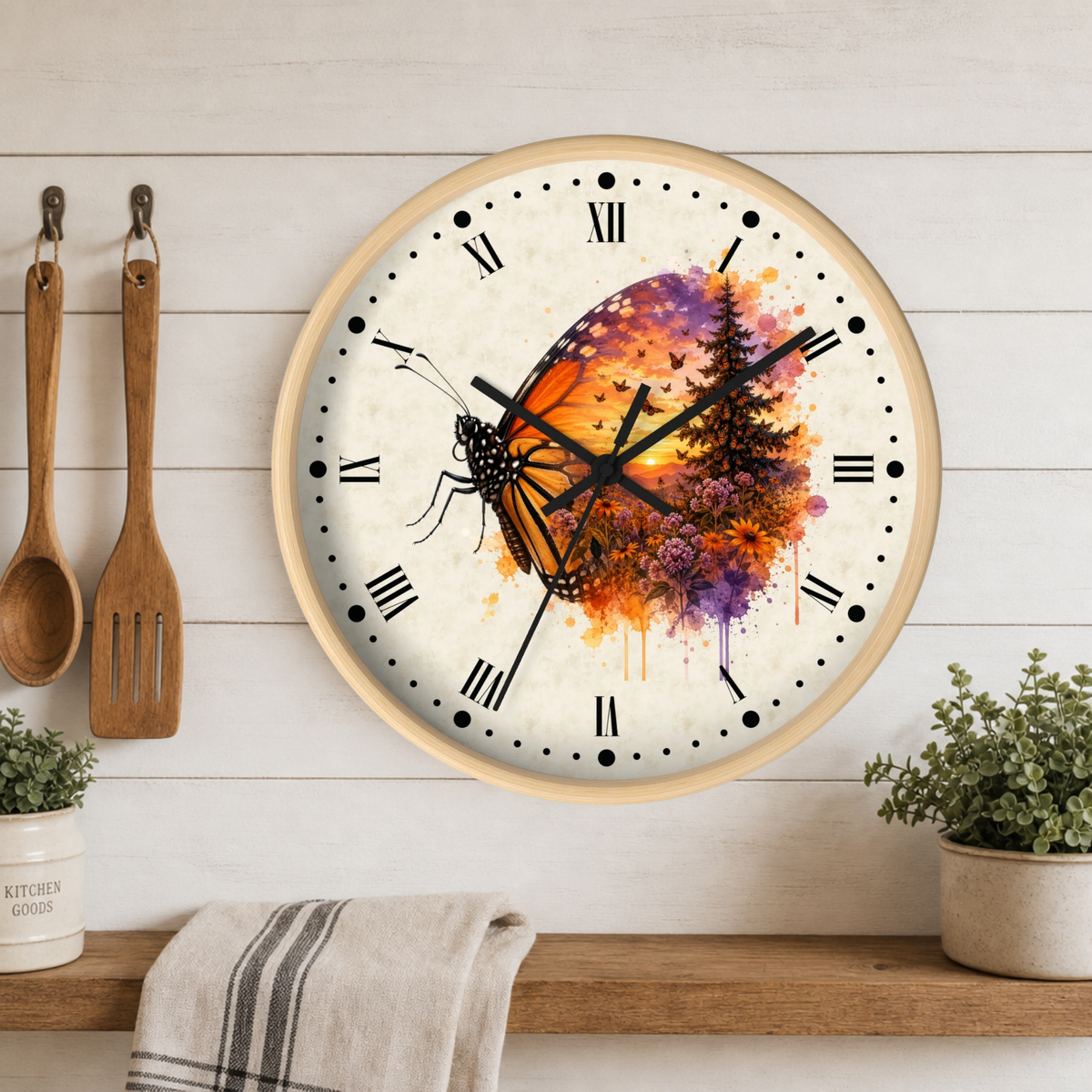 A round wall clock with a light wood frame hangs on a white wood plank wall above a wooden shelf. The clock face features a colorful butterfly and tree watercolor design in orange, purple, and yellow on a cream background.