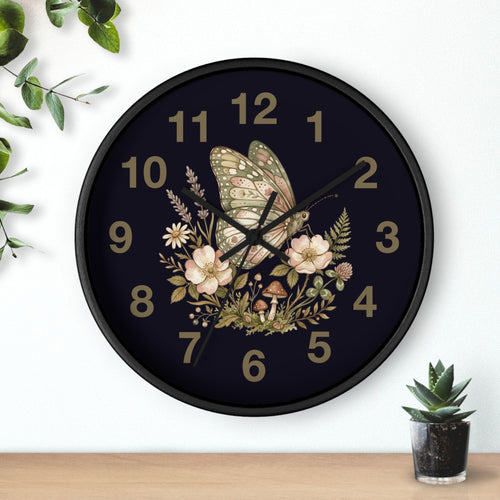 Decorative round black wall clock with butterfly and floral design on a dark background, placed on a white surface with a plant in the corner.