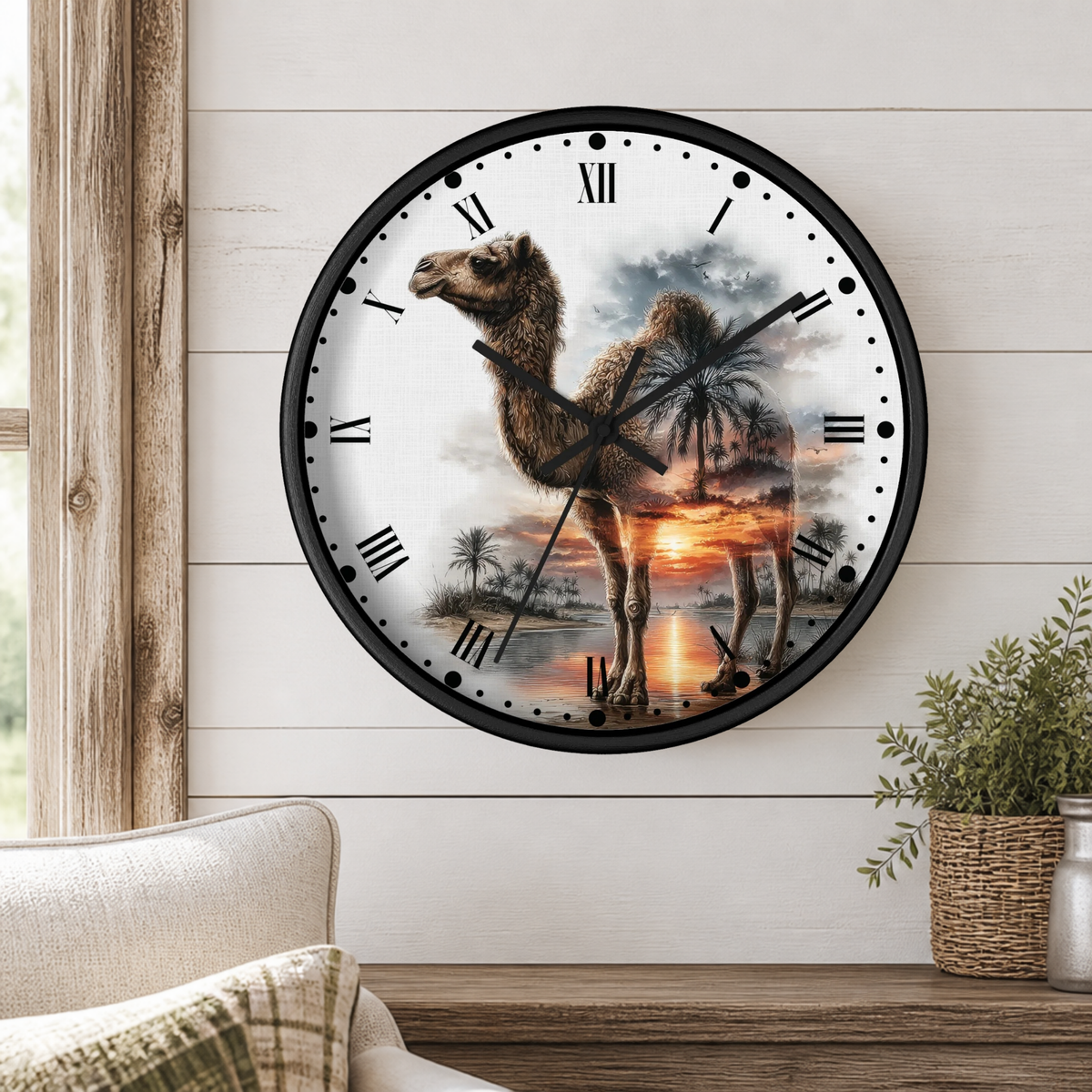 A round wall clock with a black frame hangs on a white shiplap wall above a wooden table. The clock face features a printed desert scene with a camel standing in front of a sunset over water, with black Roman numerals and hands.