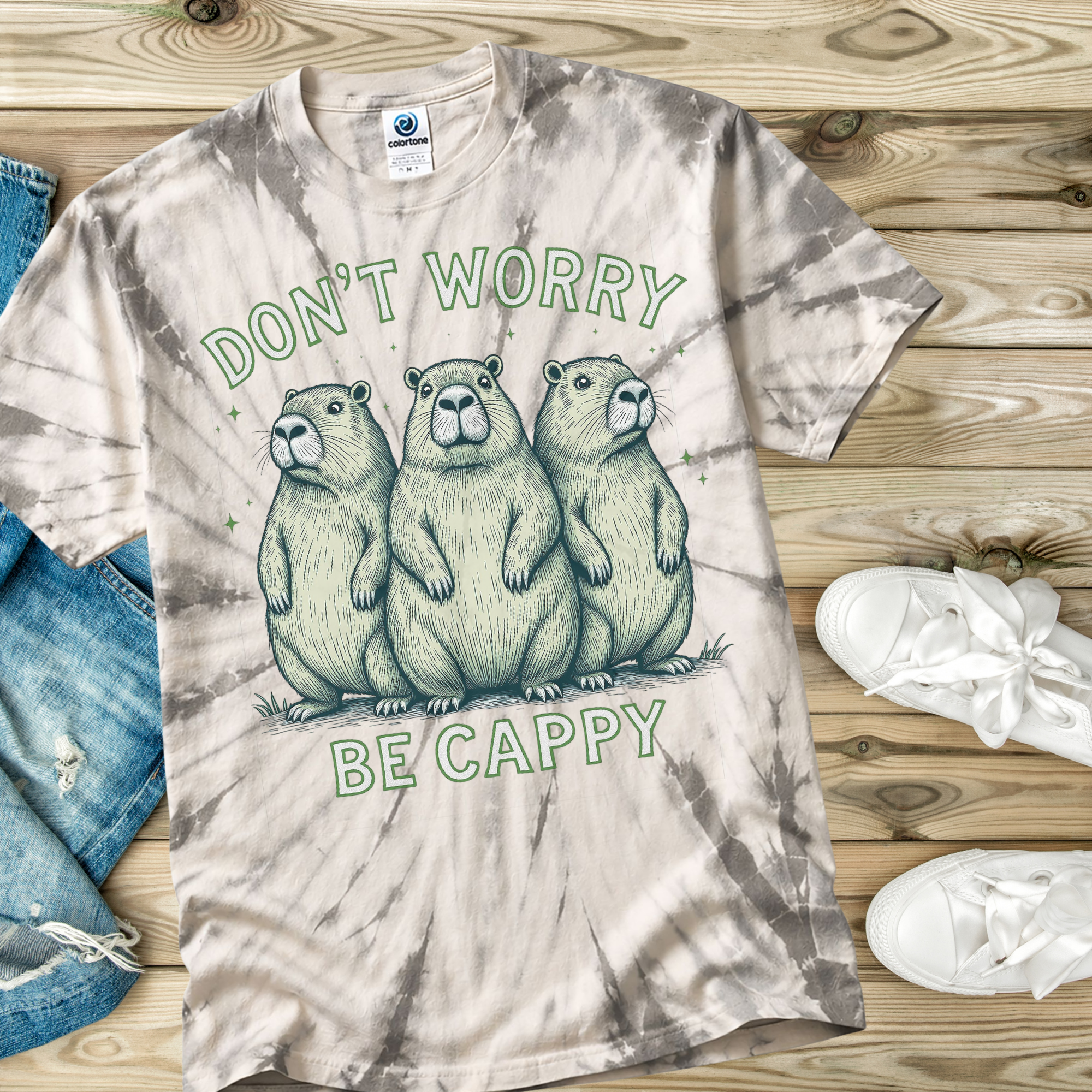 Tie-dye t-shirt with capybara design and text 'Don't Worry, Be Cappy' on a wooden surface.