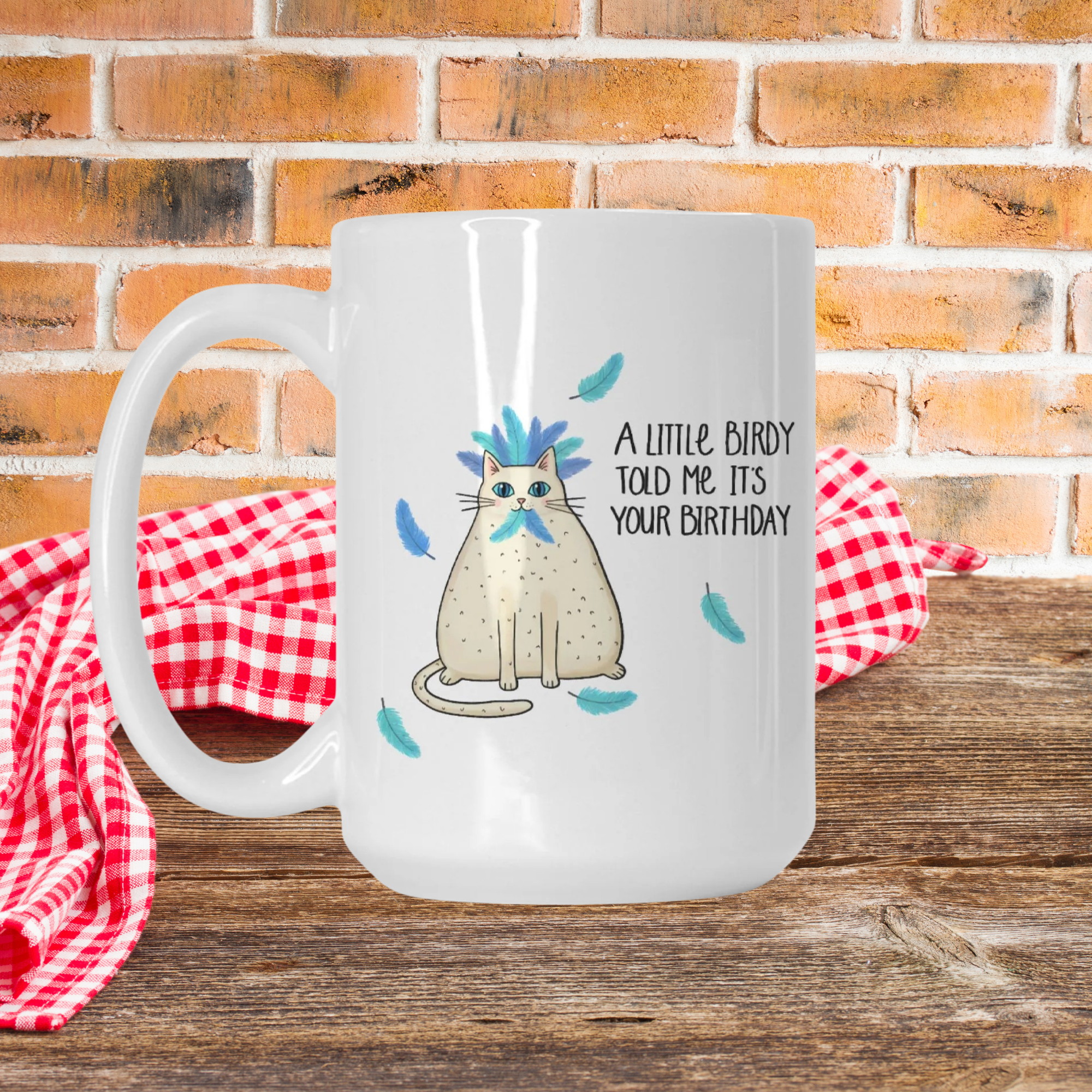 White mug with a cat graphic and text on a wooden surface with a red checkered cloth and brick wall background.