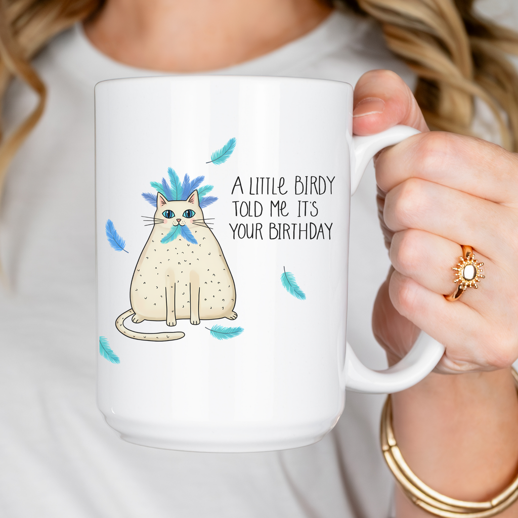 White mug with a cat illustration and birthday message, held by a person.