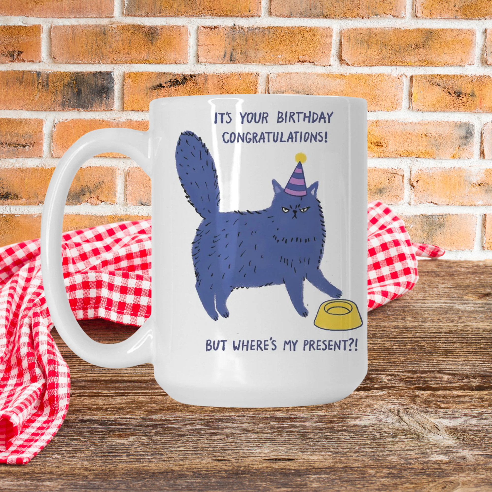 Mug with a blue cat illustration and birthday text on a wooden surface with a brick wall background