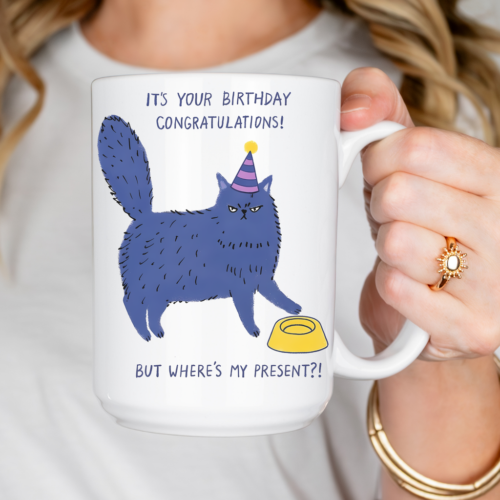 Person holding a mug with a blue cat illustration and birthday text