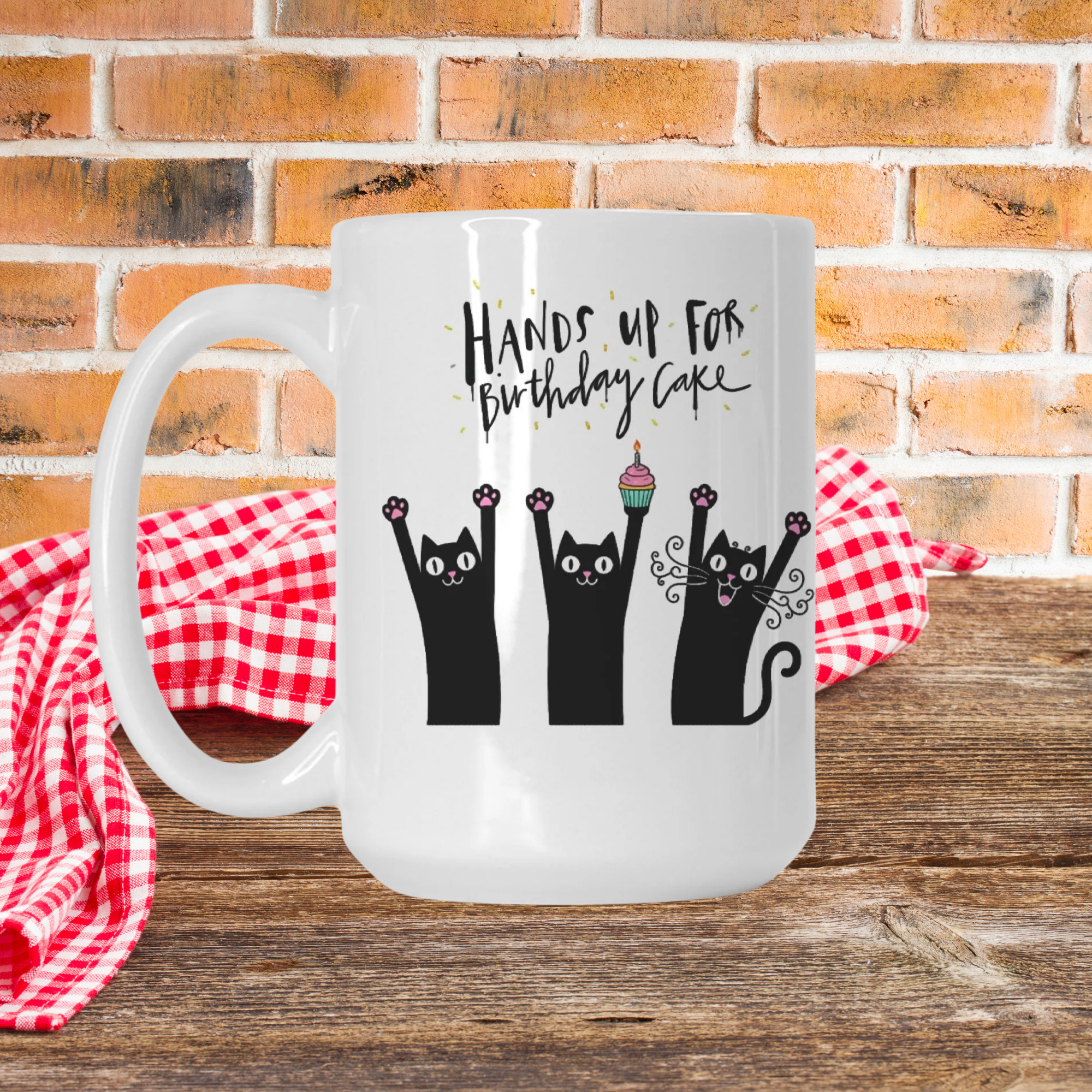 White mug with black cat design and text 'Hands up for Birthday Cake' on a wooden picnic table with a red checkered cloth and brick wall background.