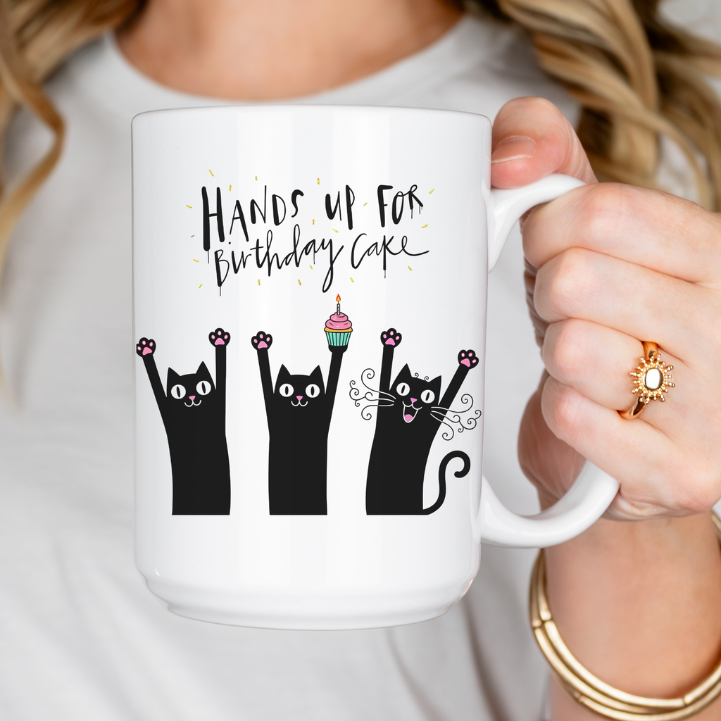 Person holding a white mug with black cats and birthday cake text design.