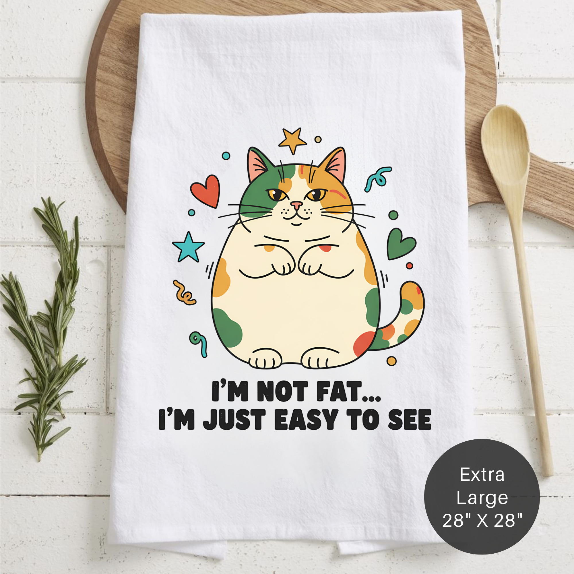White tea  towel with a colorful cat illustration and text 'I'm Not Fat... I'm Just Easy To See' on a wooden surface.
