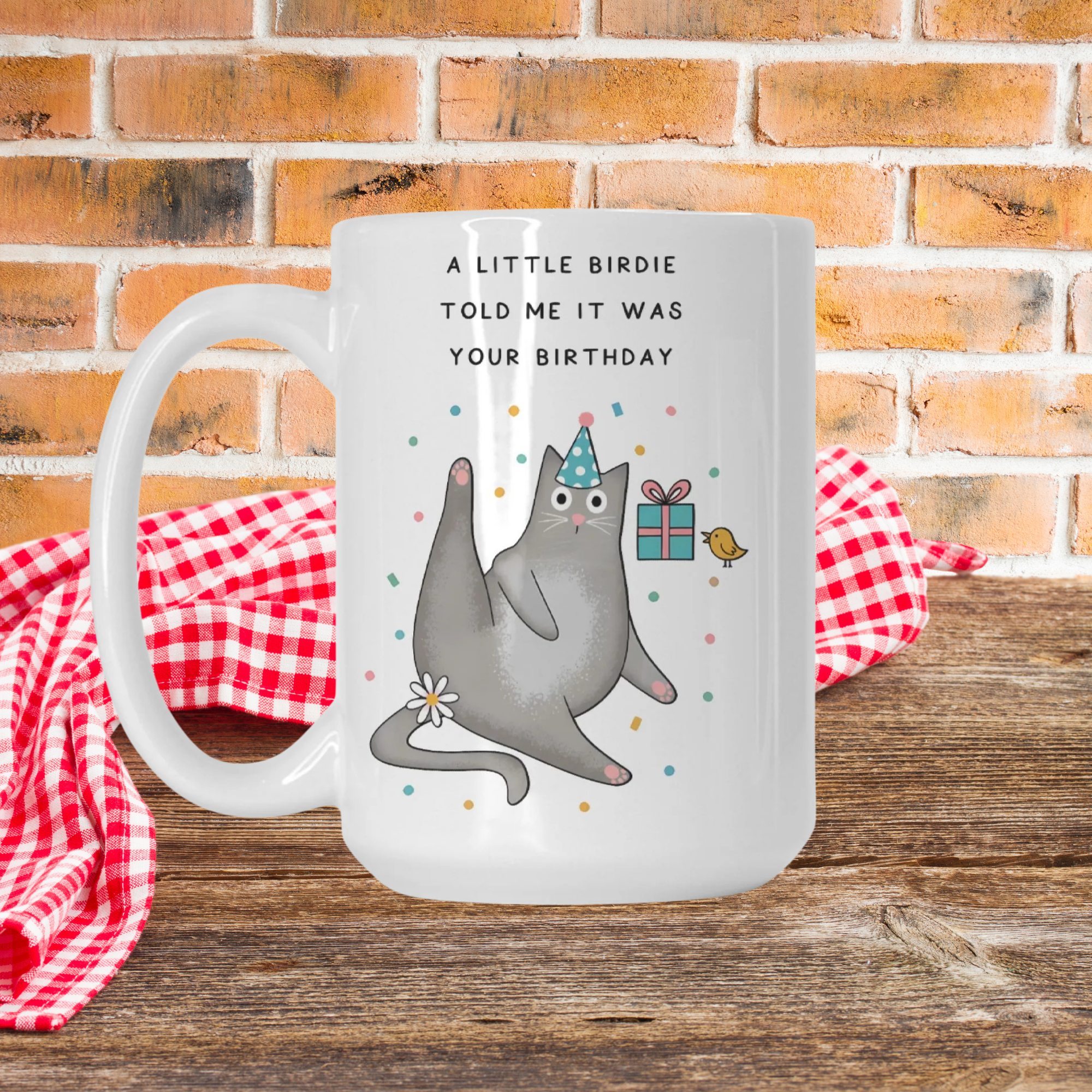 White mug with a cat illustration and birthday message on a wooden surface with a red checkered cloth and brick wall background.
