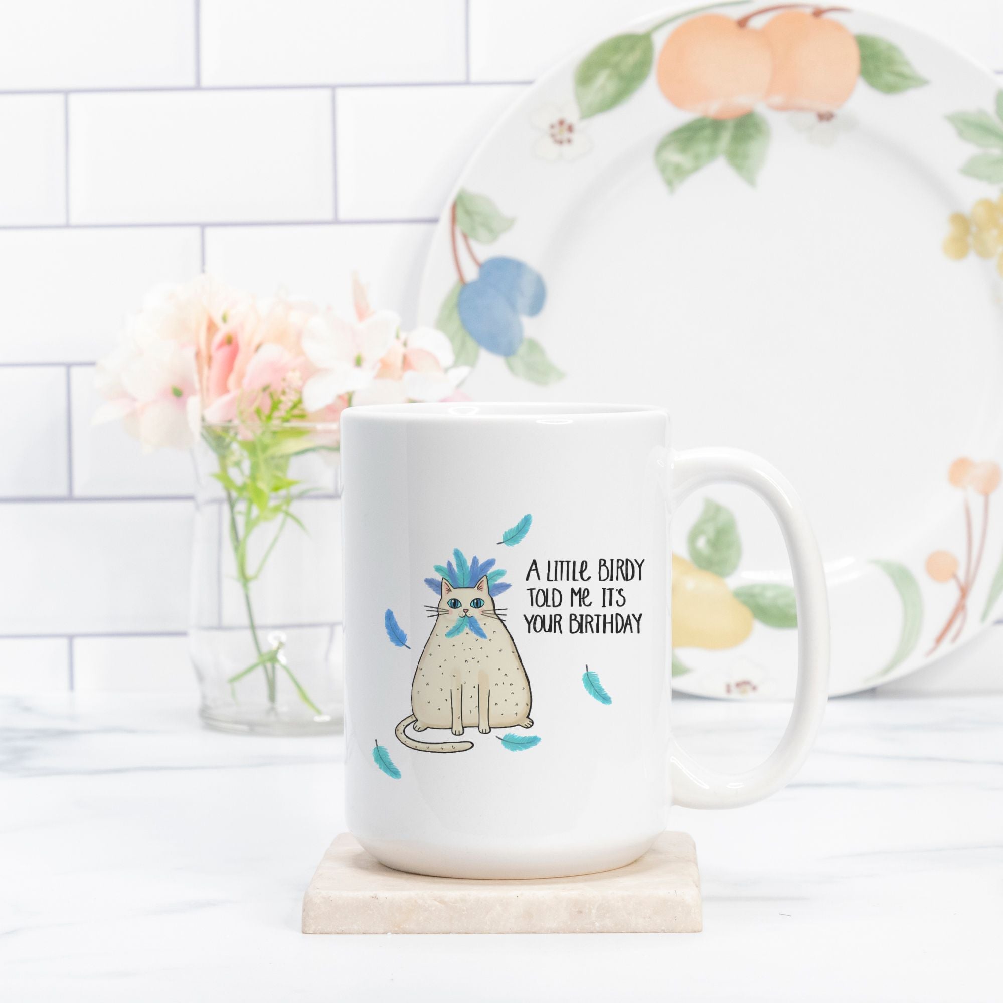 White mug with a cat illustration and text, placed on a white surface with a floral plate in the background.