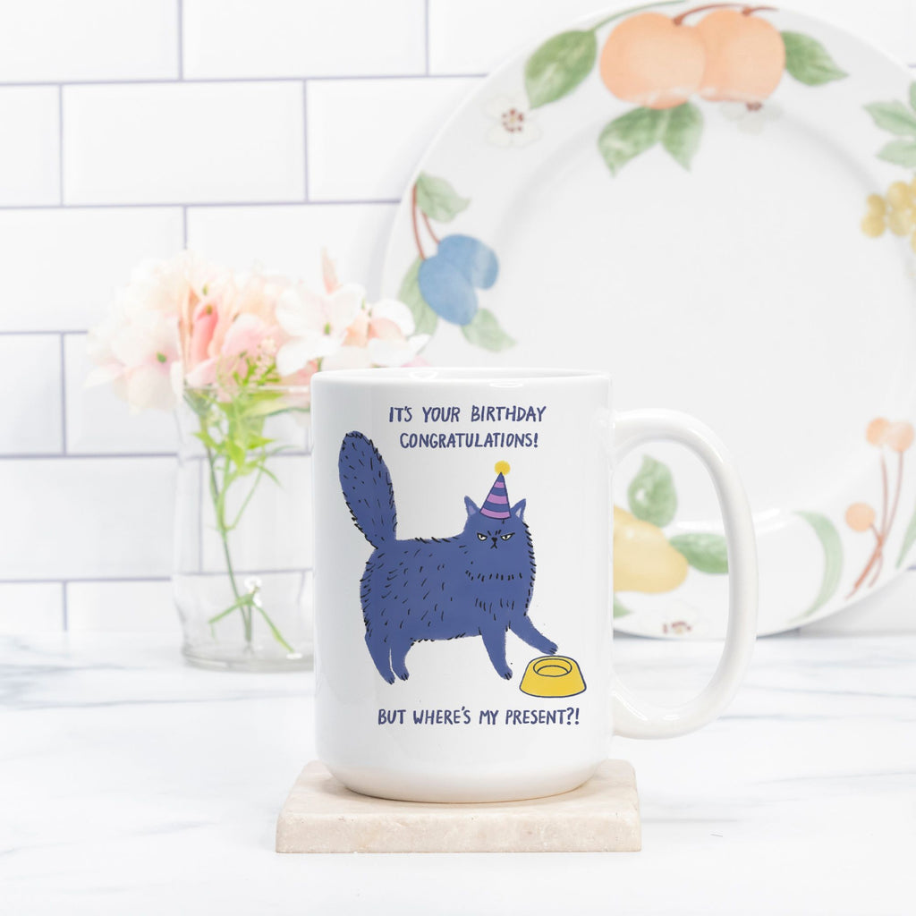 White mug with a blue cat illustration and birthday message, placed on a white surface with a floral plate in the background.