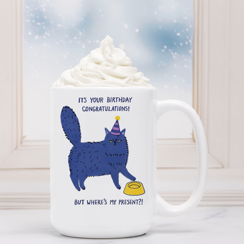 White mug with a blue cat illustration and text, topped with whipped cream against a snowy window background.