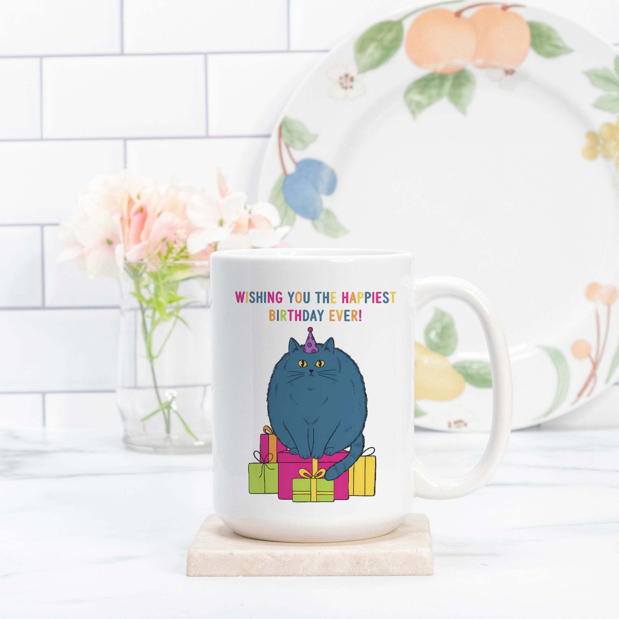 White mug with a blue cat graphic and birthday message, placed on a white surface with a decorative plate in the background.