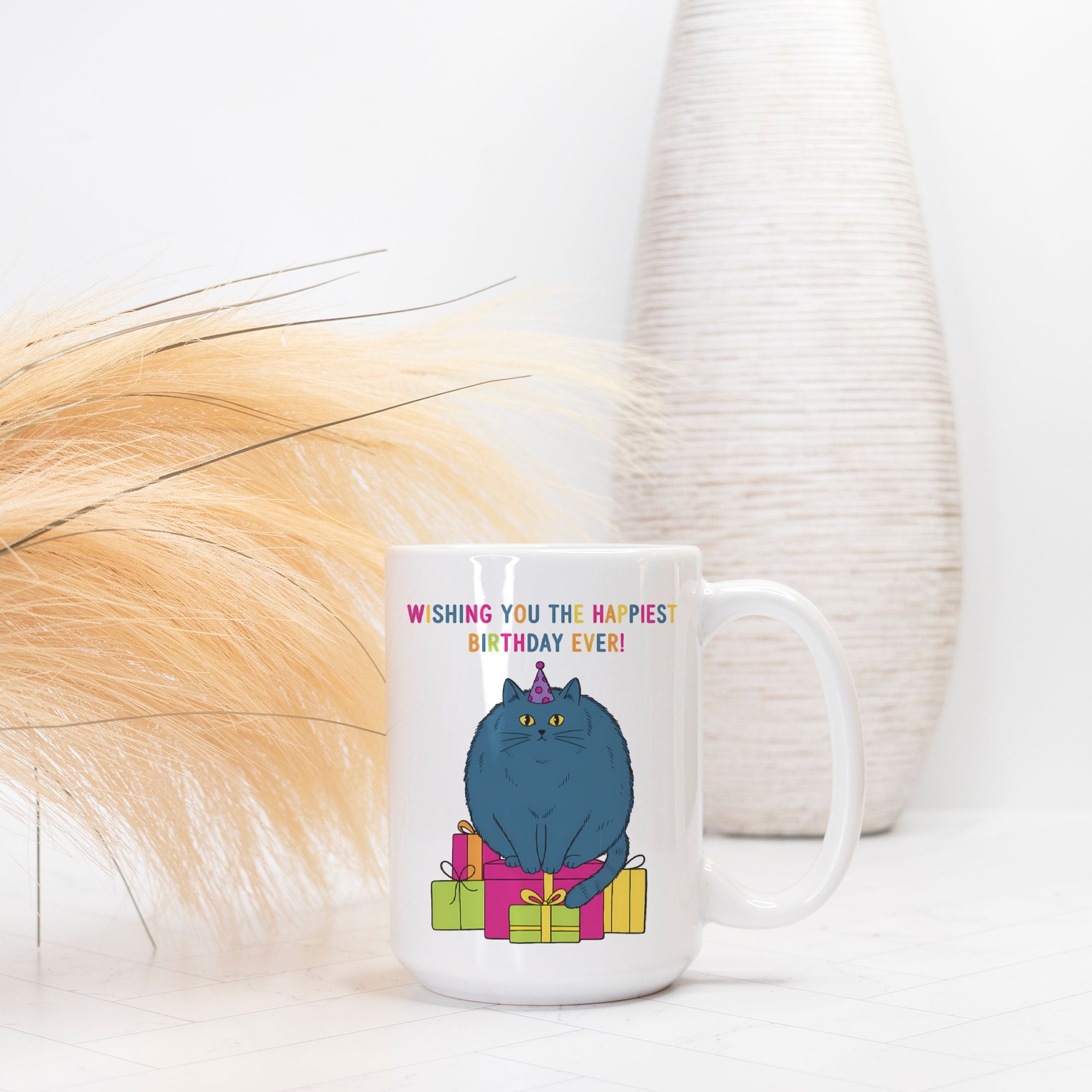 White mug with a blue cat illustration and birthday text, placed on a white surface with decorative elements.